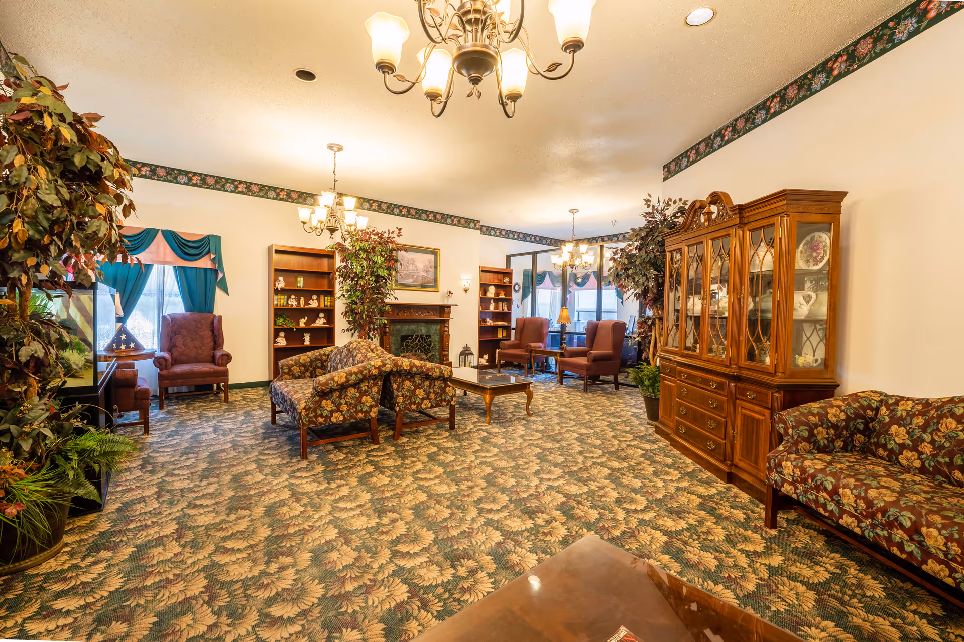 A cozy common room in a senior living facility featuring floral carpet, upholstered sofas and armchairs, a wooden china cabinet, bookshelves, a fireplace, and potted plants.