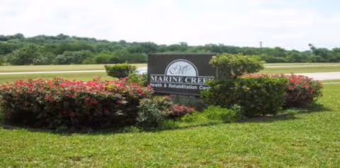 A landscaped outdoor area with green grass, bushes, and red flowers surrounding a sign that reads 'Marine Creek Health & Rehabilitation Center' with trees and a road in the background under a partly cloudy sky.