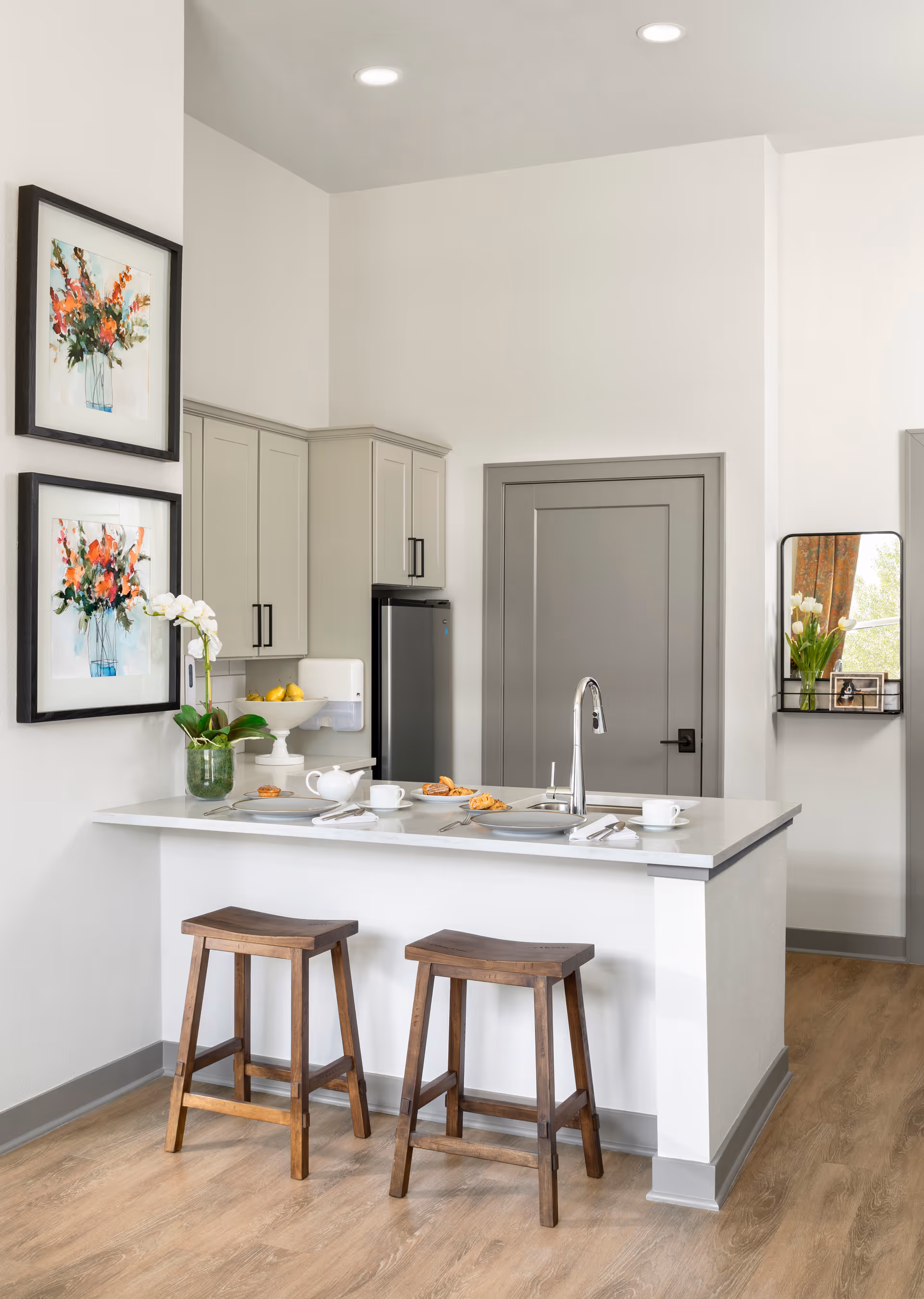 Bright modern kitchen with a white island, two wooden stools, sink and place settings, gray cabinets and framed floral artwork on the wall.