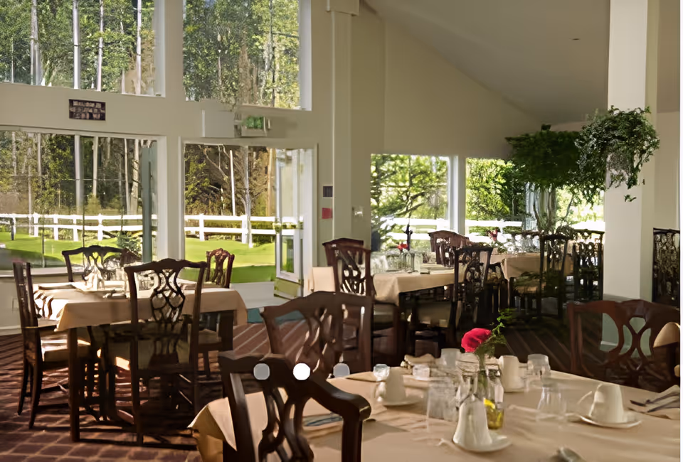 A bright dining room with multiple tables covered in beige tablecloths, set with glasses, cups, and silverware. The room has large windows and glass doors that provide a view of a green outdoor area with trees and a white fence. There are wooden chairs with decorative backs around the tables, and a hanging plant adds greenery inside the room.