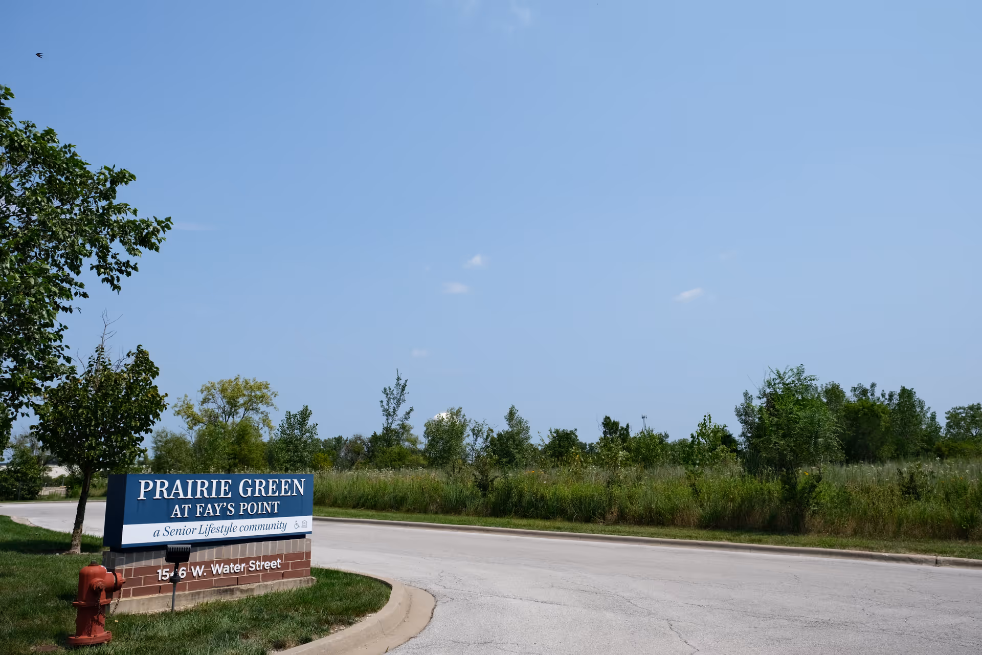 Outdoor scene showing a road curving to the left with a sign on the grass beside it that reads 'PRAIRIE GREEN AT FAY'S POINT a Senior Lifestyle community 1546 W. Water Street'. There are trees and tall grass in the background under a clear blue sky.