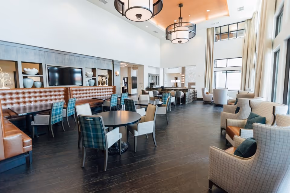 A spacious and modern dining area with high ceilings and large windows allowing natural light to fill the room. The space features round and rectangular tables with a mix of plaid and solid upholstered chairs, along with a long tufted leather bench along one wall. Decorative shelves with vases and art pieces are built into the wall behind the bench seating. Large pendant lights hang from the ceiling, and there are comfortable armchairs with pillows near the windows.