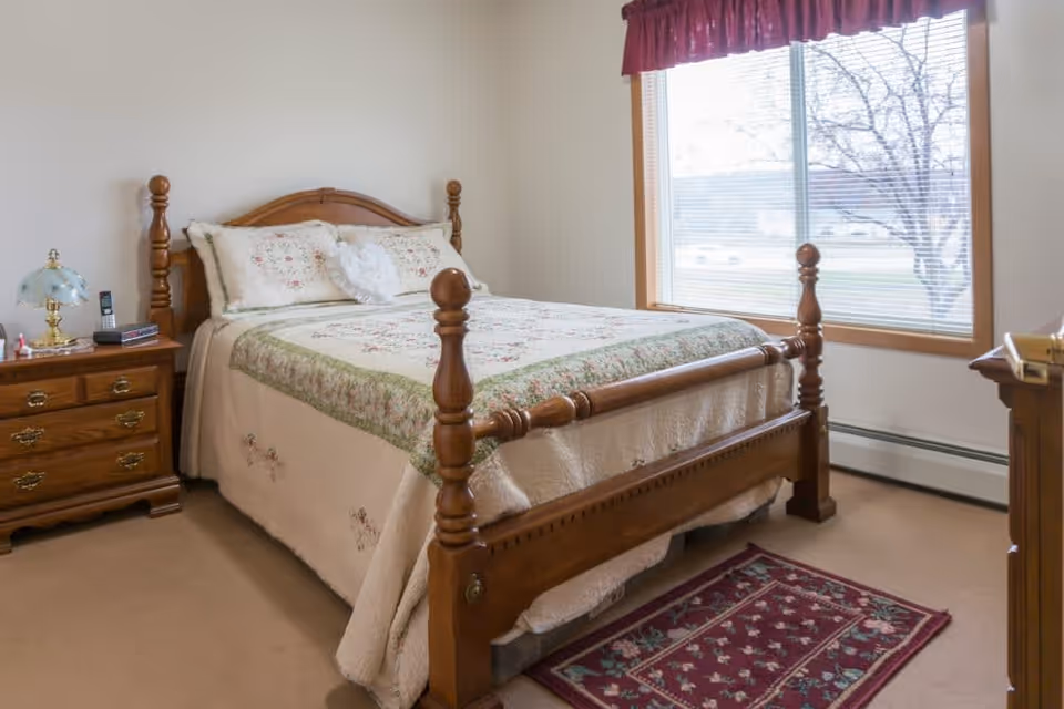 Sunlit bedroom with a wooden four-poster bed dressed in a floral quilt, a nightstand with a lamp and phone, and a large window with blinds.