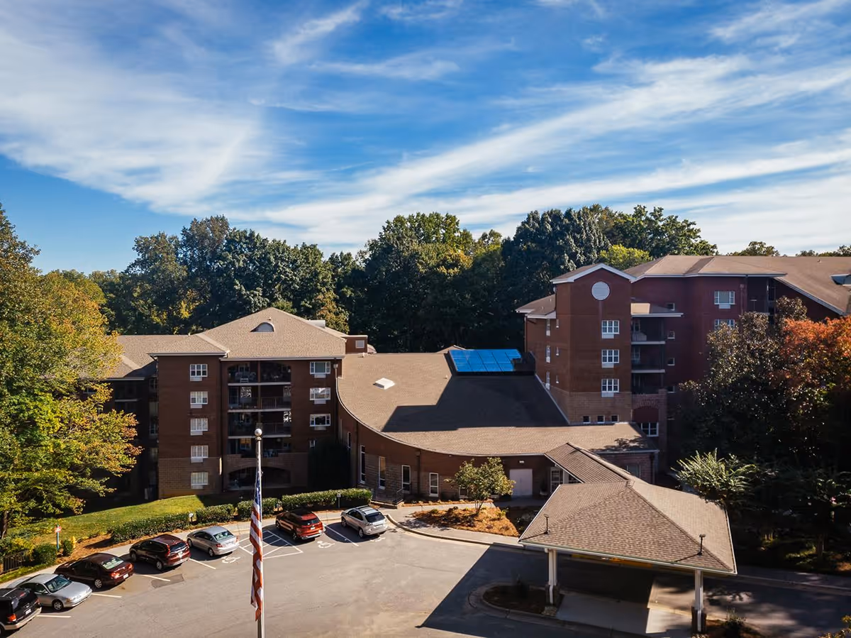 Exterior view of a multi-story brick senior living facility surrounded by trees under a blue sky with scattered clouds. Several cars are parked in the parking lot in front of the building, and an American flag is visible near the entrance.
