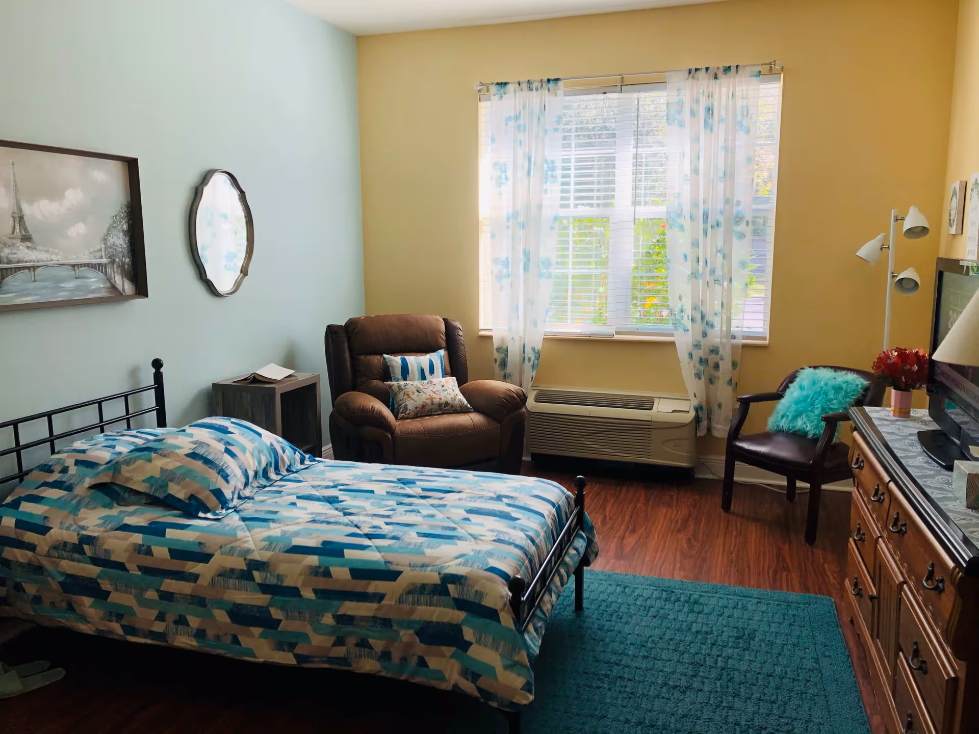 A bright furnished bedroom with a single bed, recliner, dresser and a window with floral curtains.