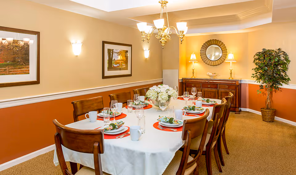 A warmly lit dining room with a long table set for eight people. The table is covered with a white tablecloth and has red placemats, white plates, glasses, mugs, and napkins with decorative napkin rings. The room features beige and orange walls, two framed pictures, a chandelier, wall sconces, a wooden sideboard with two lamps and a decorative mirror above it, and a potted plant in the corner.