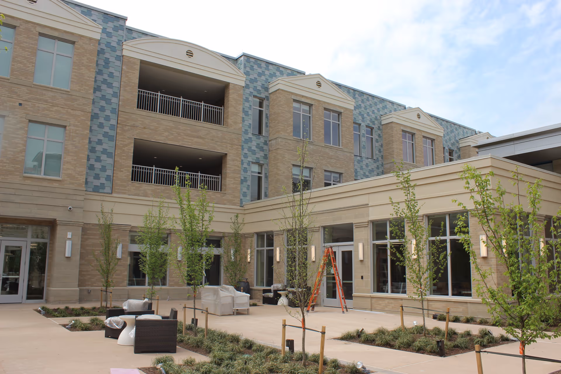 Courtyard and entrance of a multi-story brick building with balconies, young trees, and outdoor seating.