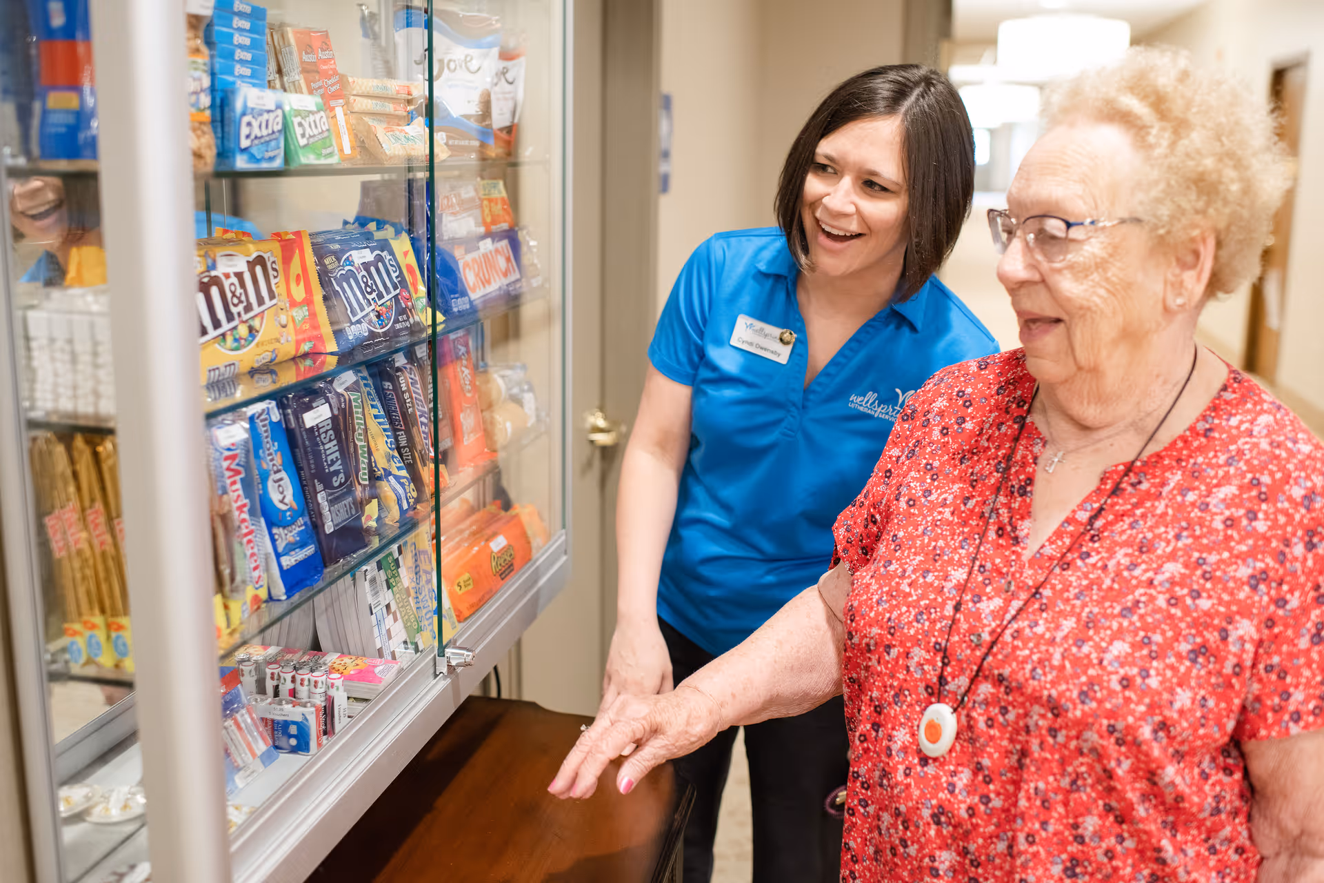 An elderly woman in a red floral shirt and a staff member in a blue Wellspring Lutheran Services shirt stand next to a glass display case filled with various candy and snack items. The staff member is smiling and engaging with the elderly woman in a hallway setting.