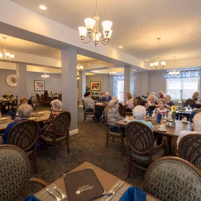 A dining room in a senior living facility with multiple elderly residents seated at round tables, engaged in conversation and dining. The room is well-lit with chandeliers, decorated with framed artwork on the walls, and has large windows with patterned curtains allowing natural light to enter.
