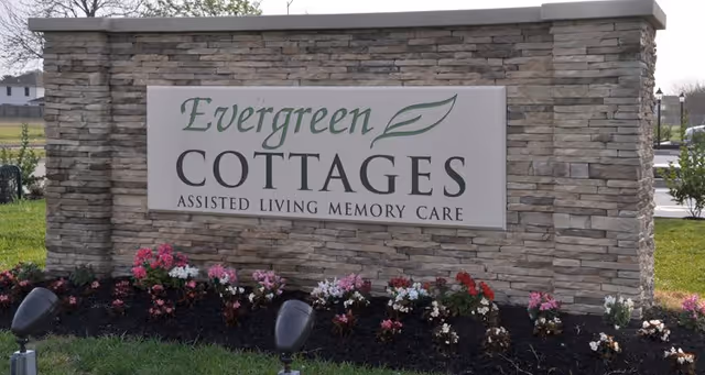 Stone monument sign for Evergreen Cottages Assisted Living Memory Care with flowers planted at the base and grass surrounding it.