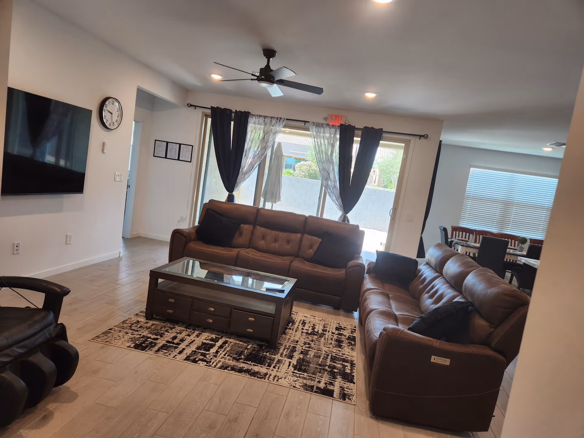 A cozy living room with two brown leather sofas facing a glass-top coffee table on a patterned rug. A large flat-screen TV is mounted on the wall to the left, and a ceiling fan with lights is above. Large sliding glass doors with dark curtains lead to an outdoor area, and a dining area with a table and chairs is visible in the background.
