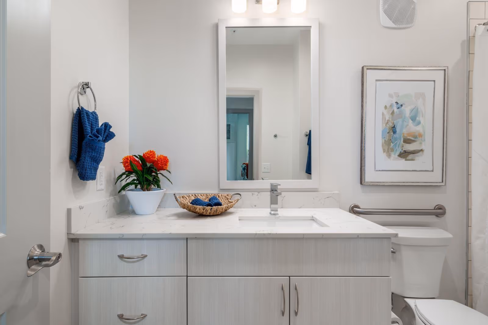 Modern white bathroom featuring a marble-top vanity with sink and mirror, blue towels, a potted plant, and a toilet with grab bar.