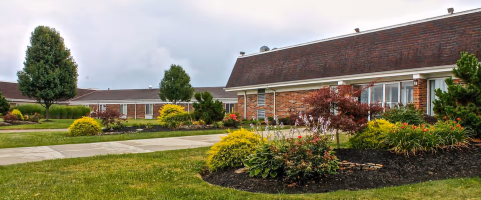 Exterior view of Beckley Healthcare Center showing a single-story brick building with a sloped roof, surrounded by well-maintained landscaping including green grass, bushes, small trees, and flower beds under a cloudy sky.