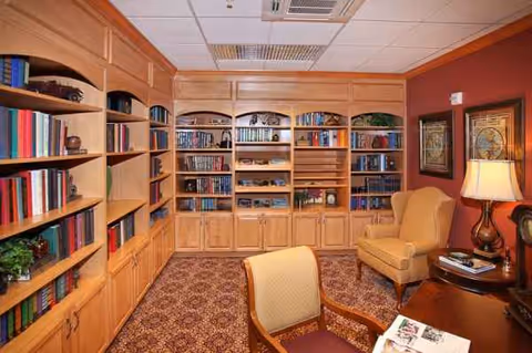 Interior library room with built-in wooden shelves, armchairs, and a reading table.