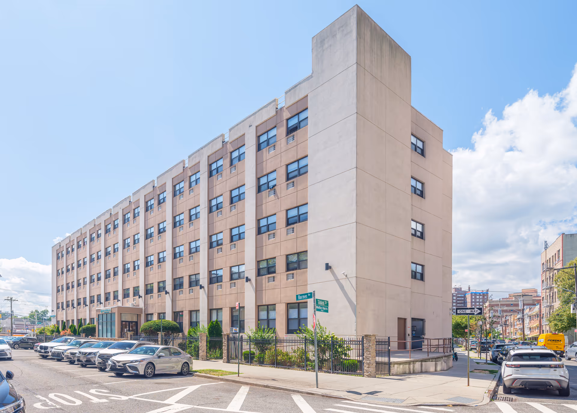 Exterior view of a multi-story beige building on a sunny day with several parked cars along the street. The building has many windows and is located at the corner of Barnes Avenue and West 215th Street, surrounded by a black metal fence and some greenery.