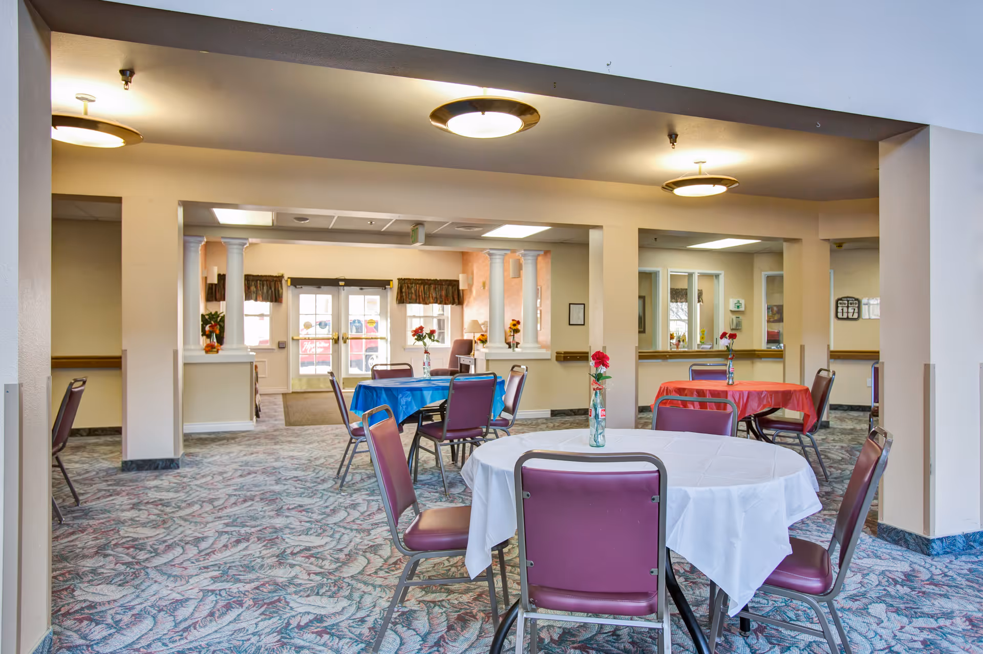 Interior view of a senior living facility dining area with round tables covered in white, blue, and red tablecloths. Each table has a vase with red flowers. The room has patterned carpet flooring, beige walls, and ceiling lights. There are multiple chairs around the tables and large windows in the background letting in natural light.