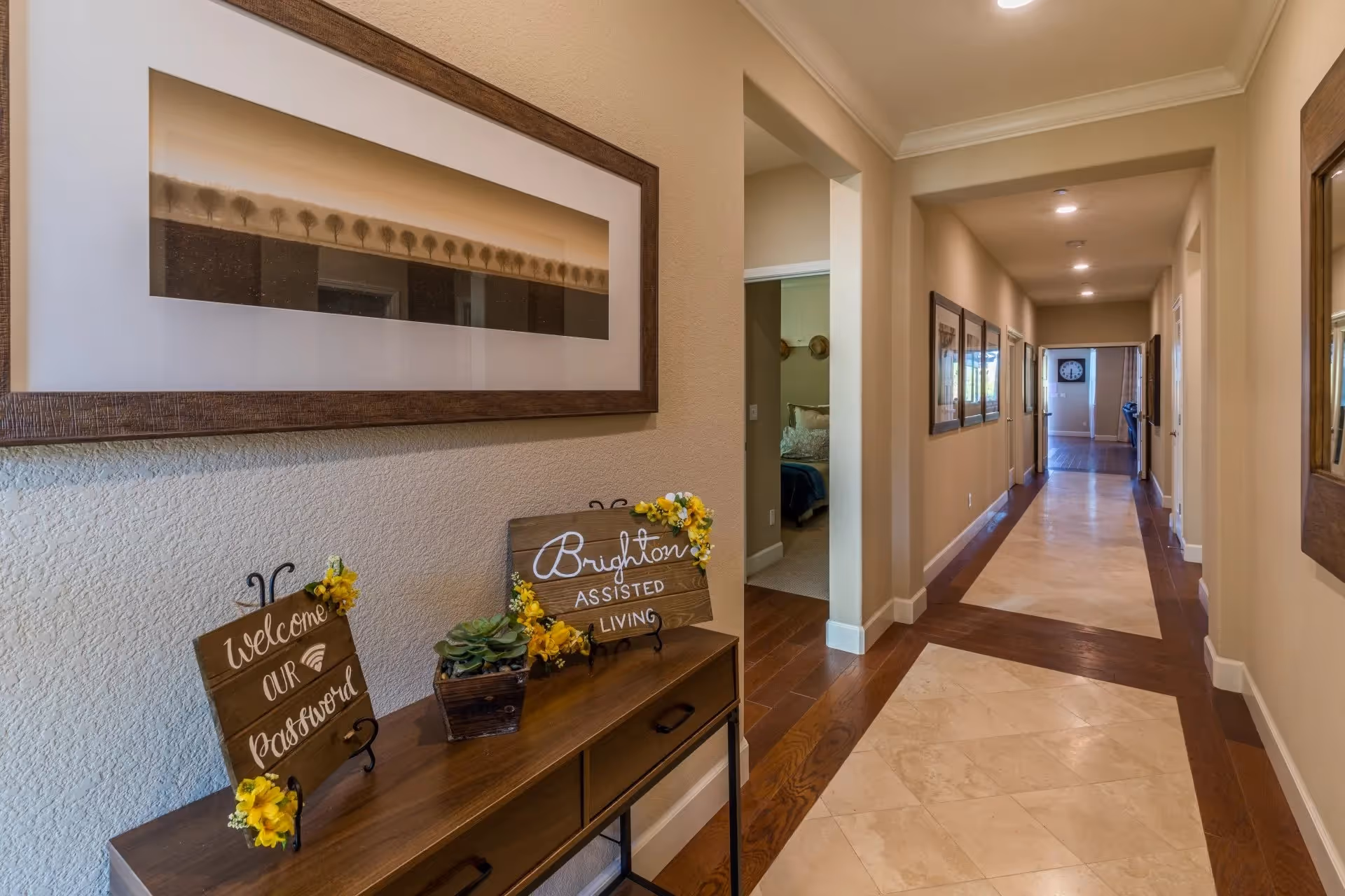 A well-lit hallway in an assisted living facility with beige walls and a tiled floor bordered by wood. On the left side, there is a wooden console table with two decorative signs, one reading 'Welcome Our Password' and the other 'Brighton Assisted Living,' along with a small potted plant. Above the table hangs a framed artwork depicting a row of trees. The hallway extends into the distance with several doorways and framed pictures on the walls.