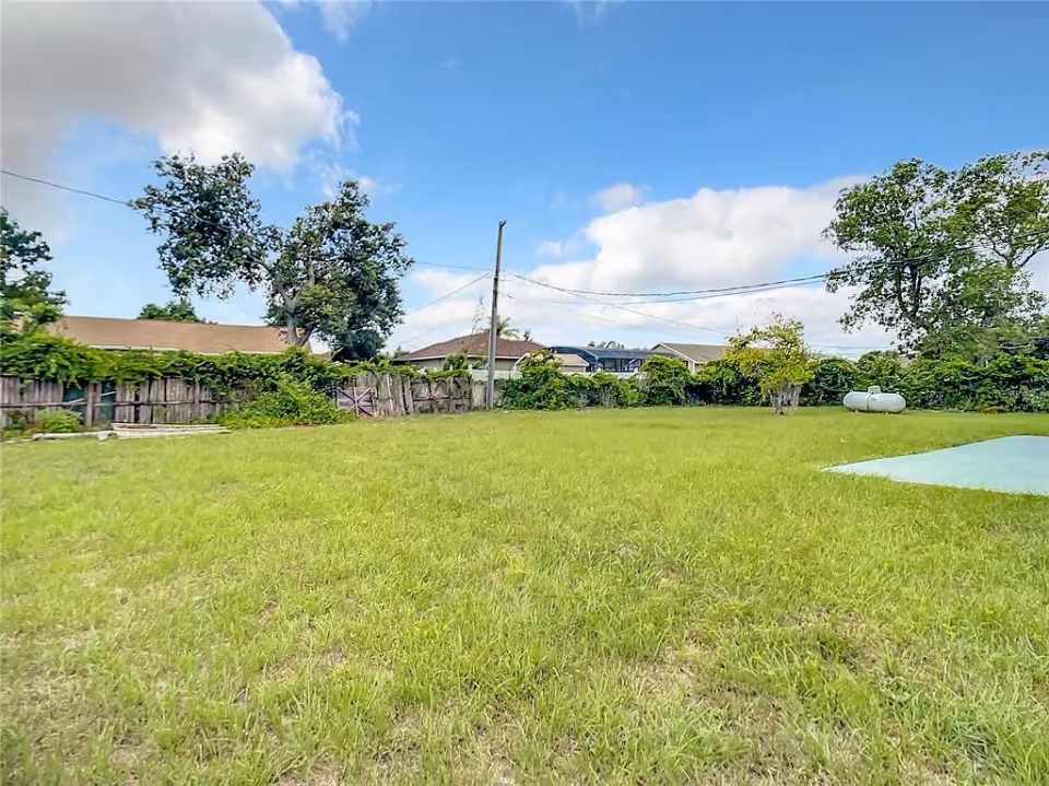 A large grassy yard with a few trees and bushes along a wooden fence. There are houses visible in the background under a partly cloudy blue sky. A propane tank is seen on the right side of the yard.