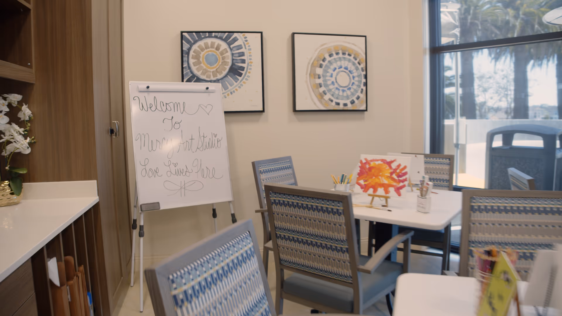 Interior view of an art studio room at Mercy Retirement & Care Center featuring a whiteboard with the message 'Welcome to Mercy Art Studio Love Lives Here', several chairs with patterned upholstery around white tables, art supplies including colored pencils and a painting, two abstract circular paintings on the wall, and a large window showing palm trees outside.