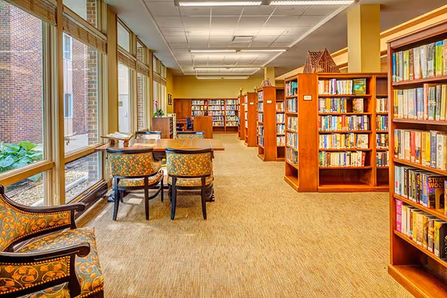 A bright and spacious library room with large windows on the left letting in natural light. There are wooden bookshelves filled with books along the right side and at the far end of the room. Several wooden tables with upholstered chairs are arranged near the windows for reading or studying. The carpeted floor and ceiling lights create a warm and inviting atmosphere.