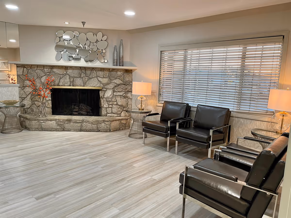 Spacious sitting room with a stone fireplace, black leather chairs, side tables and large window blinds.
