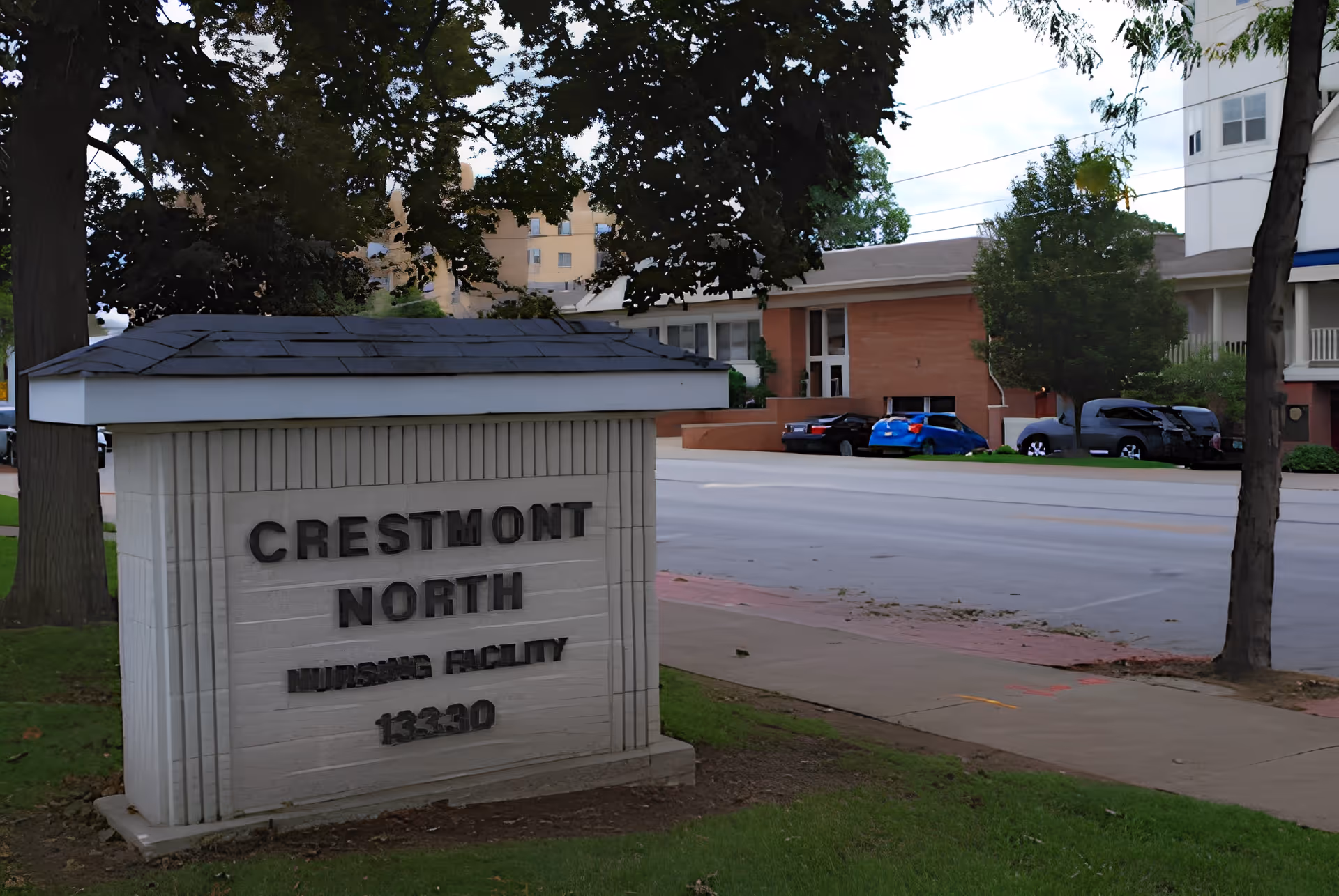 Outdoor view of a sign for Crestmont North Nursing Facility with the address 13330, situated on a grassy area near a sidewalk and street. Trees and parked cars are visible in the background along with parts of buildings.