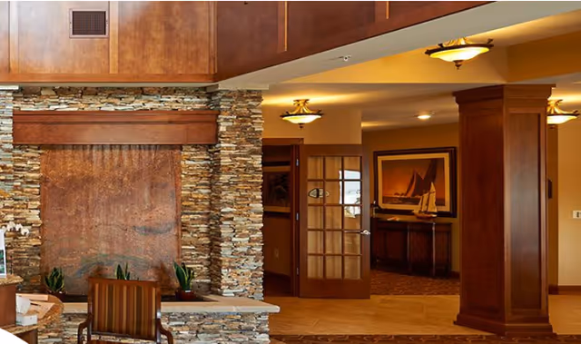 Interior view of a senior living facility featuring a stone and wood accent wall with a built-in water feature, a wooden bench with striped cushions, potted plants, warm lighting fixtures, a wooden column, and a glass-paneled door leading to another room with framed artwork and a model sailboat on a cabinet.