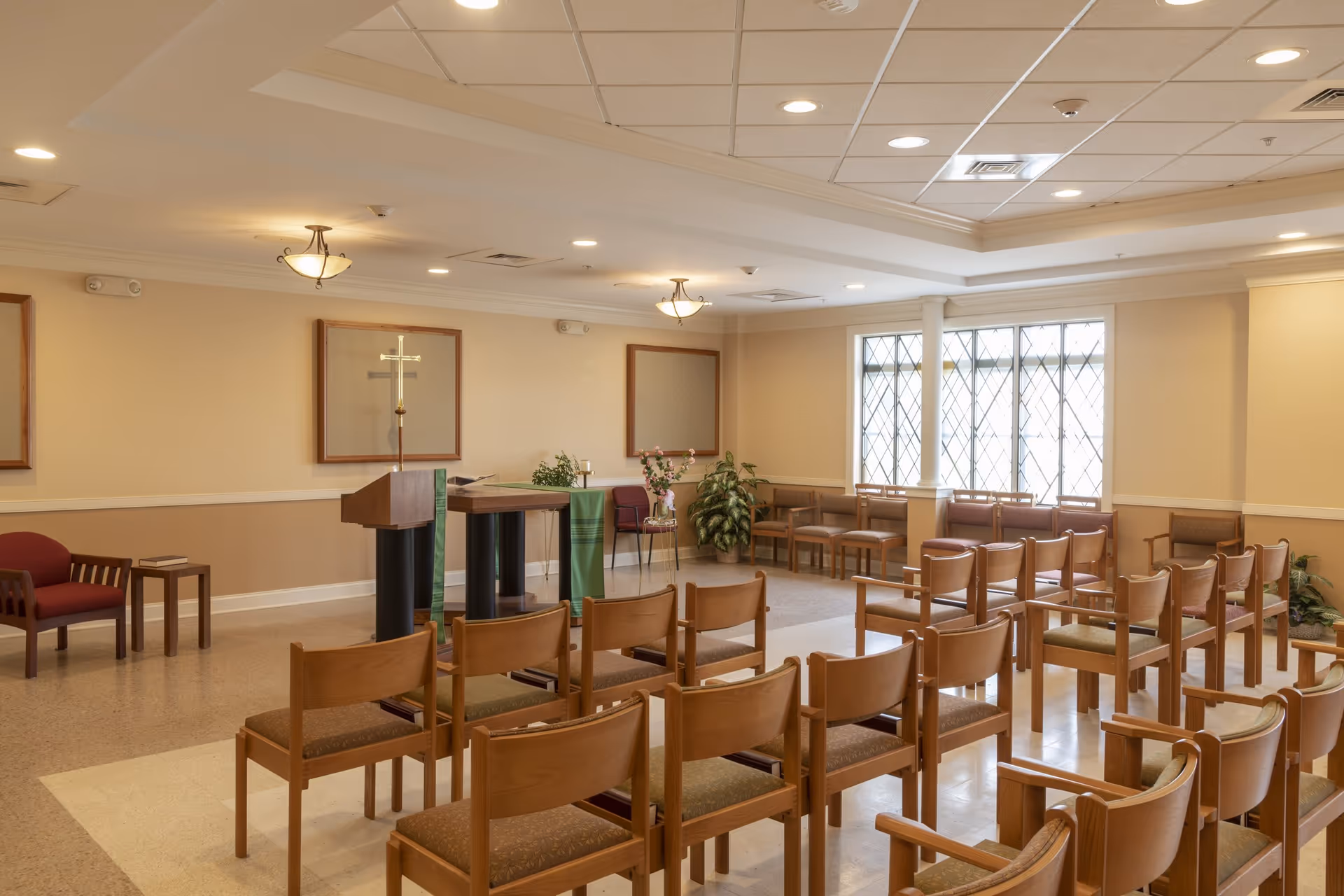 A small chapel or meeting room with rows of wooden chairs facing a podium with a cross on it. The room has beige walls, a tiled ceiling with recessed lighting, and large windows with diamond-patterned panes letting in natural light. There are a few plants and framed bulletin boards on the walls.