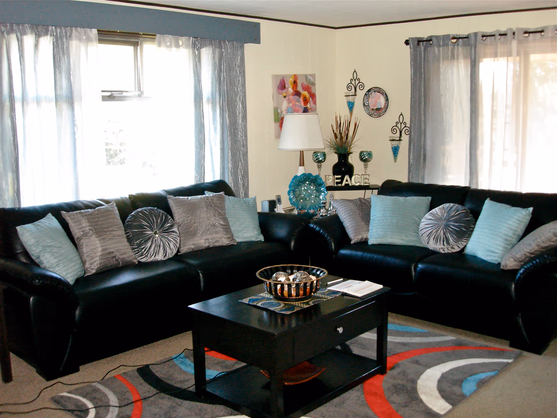 A cozy living room with two black leather sofas adorned with various blue and gray decorative pillows. A black coffee table with a decorative bowl and a book sits on a colorful patterned rug. The room has large windows with sheer curtains, and the walls are decorated with a lamp, floral artwork, wall sconces, and a decorative piece spelling 'PEACE'.