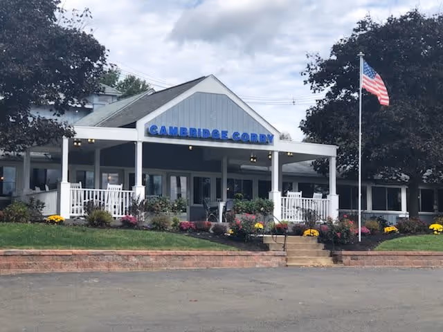 Front exterior view of Cambridge Corry Senior Living building with a porch, white railing, flowers in the garden, and an American flag on a flagpole to the right.
