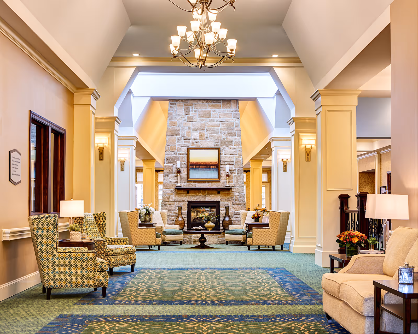 Bright, elegant communal lounge with upholstered chairs and sofas arranged around a central stone fireplace under a chandelier.