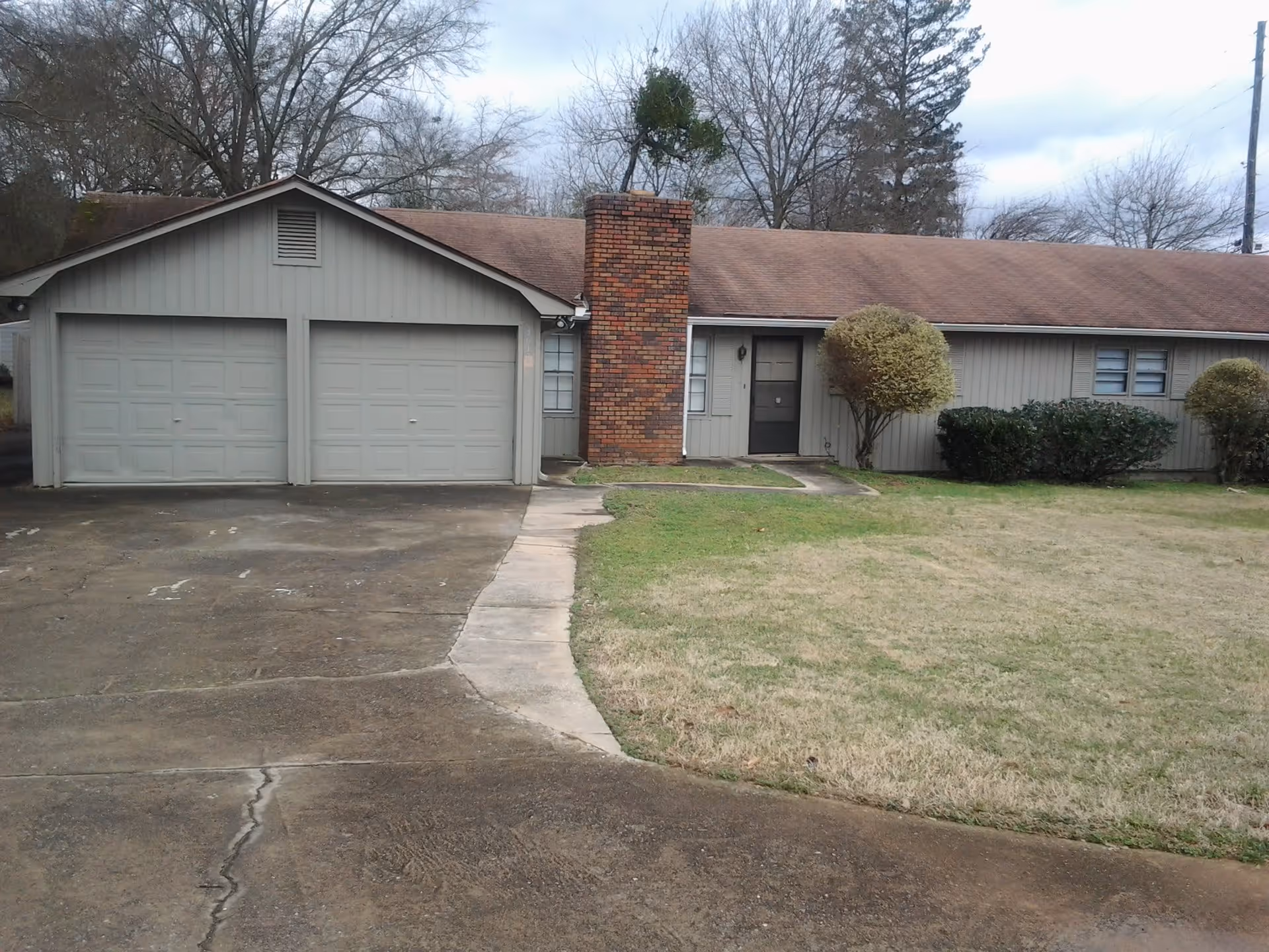 Exterior view of a single-story residential building with a double garage, a brick chimney, and a front door. The building is surrounded by a lawn with trimmed bushes and trees in the background under a cloudy sky.
