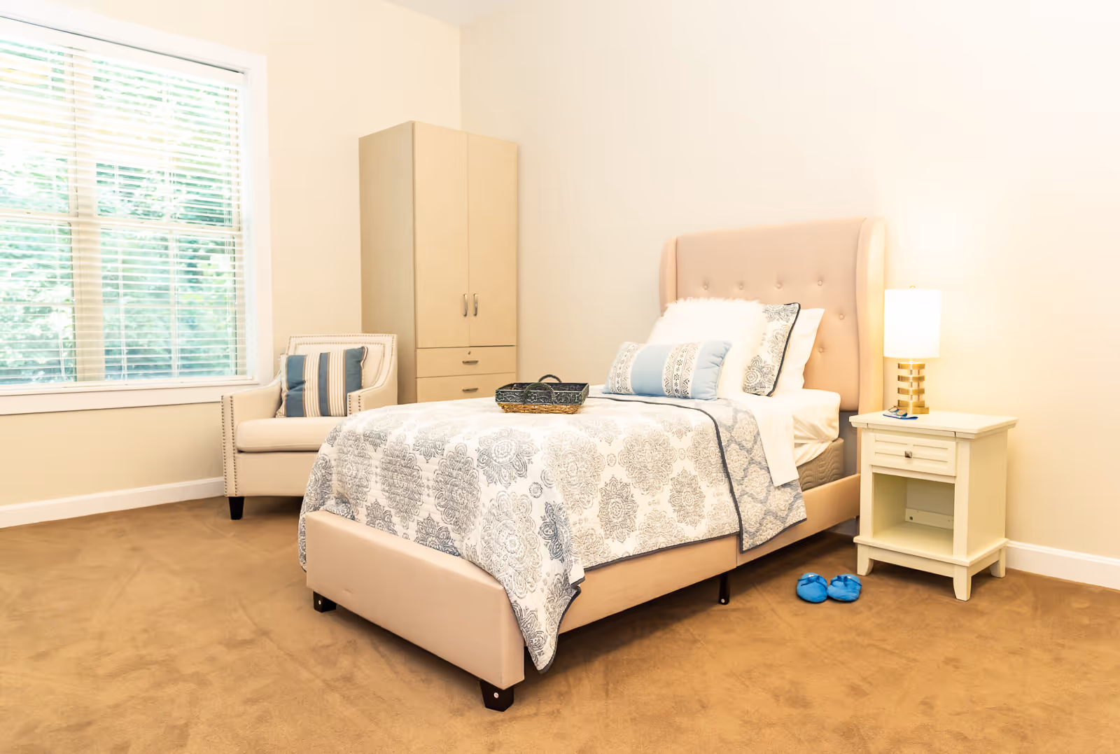 A bright bedroom with a beige upholstered bed featuring patterned bedding and multiple pillows. Next to the bed is a white nightstand with a lamp and a pair of blue slippers on the carpeted floor. A beige armchair with a striped cushion is positioned near a large window with white blinds. A tall beige wardrobe stands against the wall.
