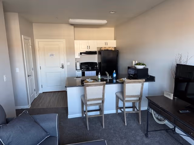 Open-plan apartment interior with a kitchen island and two bar stools, a sofa, TV, and black appliances.