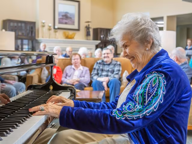 An elderly woman smiling while playing a grand piano for a seated group of seniors in a communal living room.