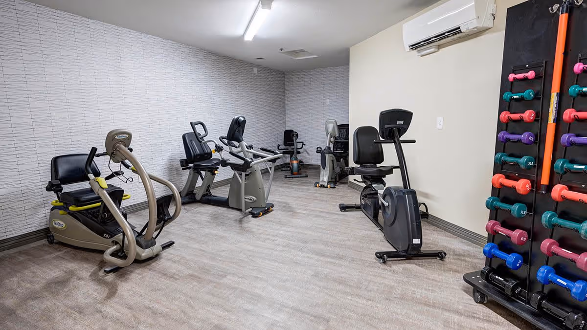 A small fitness room with several exercise machines including recumbent bikes and an elliptical trainer. There is a rack of colorful dumbbells on the right side of the room. The walls are light-colored with a textured pattern on one side, and the floor has a wood-like finish. An air conditioning unit is mounted on the wall.