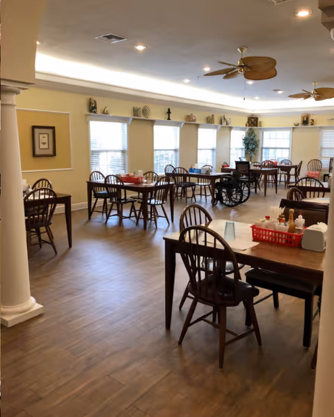 Bright dining room with multiple wooden tables and chairs, ceiling fans, windows, and a wheelchair near the back.