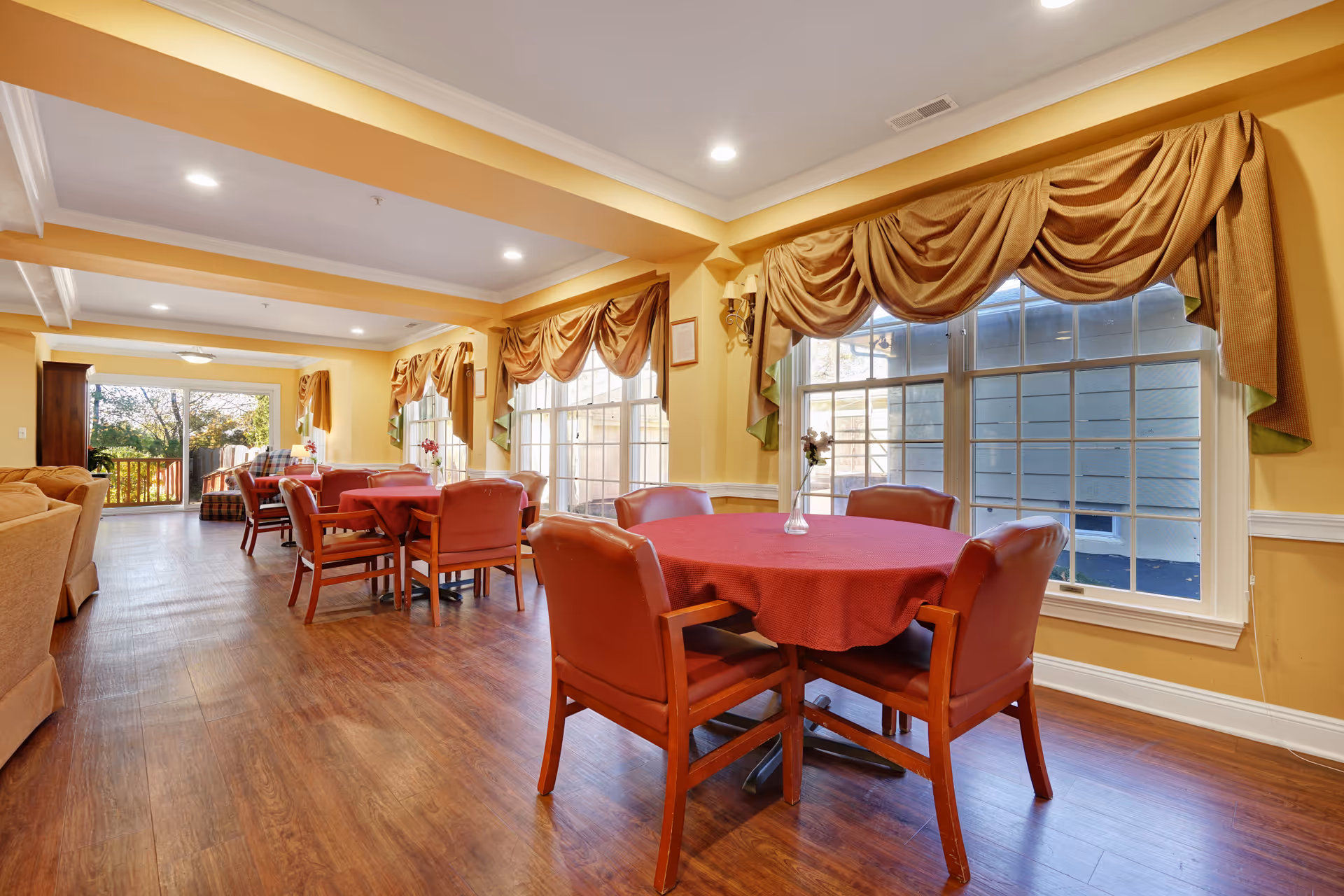 A bright and spacious common area with several round tables covered in red tablecloths, each surrounded by four wooden chairs with red cushions. Large windows with decorative valances let in natural light, and the room features warm yellow walls and wood flooring. In the background, there is a seating area with beige sofas and a sliding glass door leading outside.