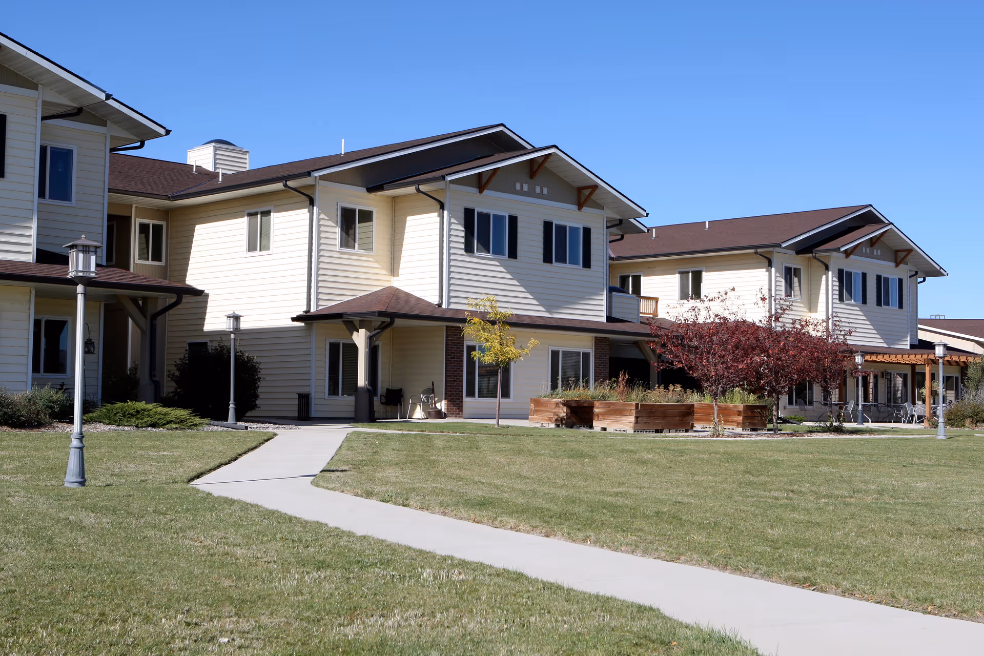Exterior view of a two-story senior living facility building with beige siding and brown roofs, surrounded by a well-maintained lawn, paved walkways, lamp posts, and raised garden beds with small trees and shrubs under a clear blue sky.