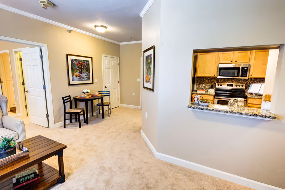 Carpeted apartment interior showing a small dining table and chairs, a living area, and a pass-through window into a kitchen with granite counter and stainless appliances.