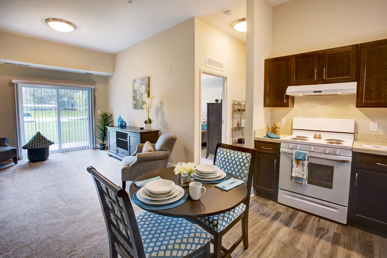 A bright and cozy living space in a senior living facility featuring a small round dining table set for two with blue patterned chairs, a kitchen area with dark wood cabinets and a white stove, a comfortable armchair, a TV stand with decorative items, and a large sliding glass door with vertical blinds leading to an outdoor view.