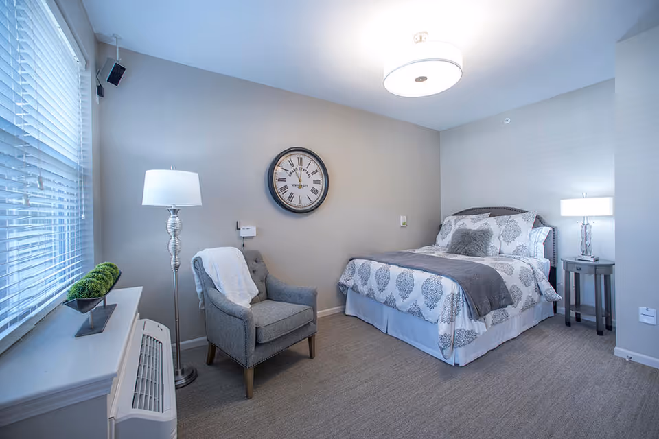 A neatly arranged bedroom featuring a bed with patterned bedding and multiple pillows, a gray upholstered armchair with a white throw draped over it, a floor lamp, a round wall clock, and a bedside table with a lamp. The room has light-colored walls and carpeted flooring, with a window covered by white blinds.