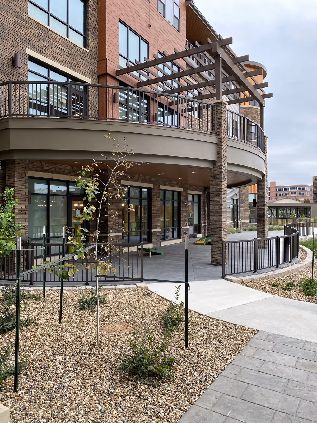 Outdoor view of a modern retirement community building with large windows, a covered patio area with railings, a small tree and shrubs planted in a gravel bed, and a paved walkway leading to the entrance.