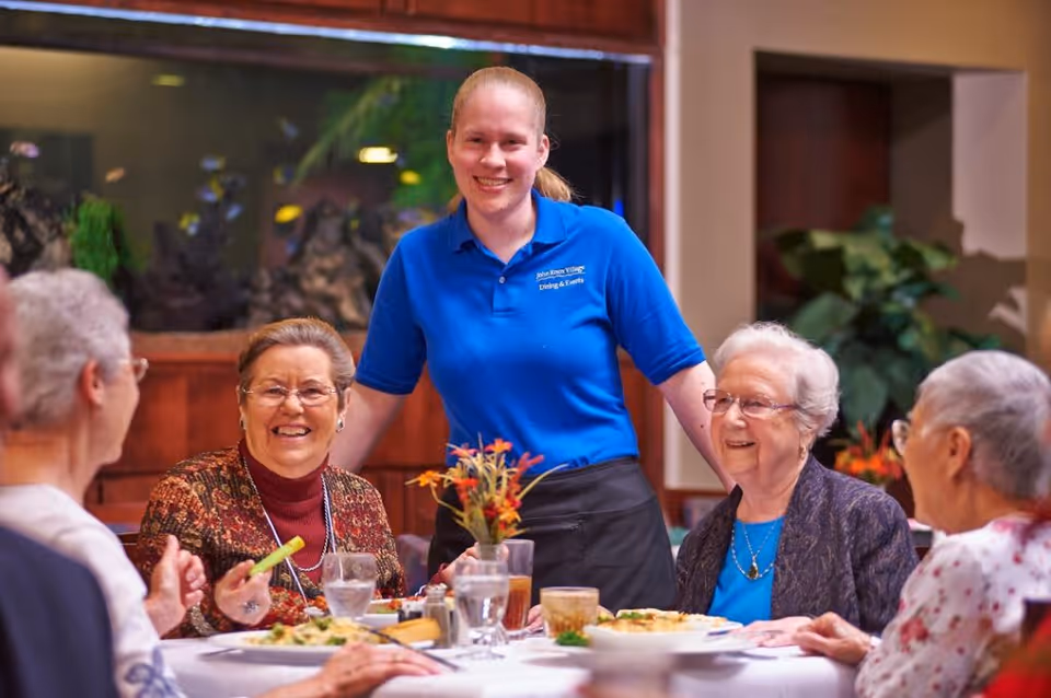 A smiling staff member in a blue John Knox Village Dining & Events polo shirt stands behind a table where four elderly women are seated and enjoying a meal together in a dining room setting with a large aquarium in the background.