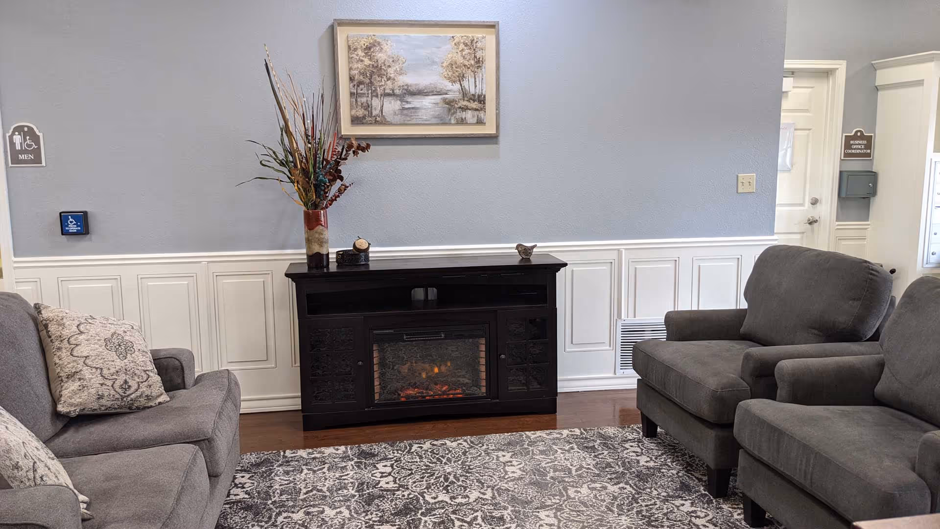 A cozy seating area with two gray armchairs and a gray sofa arranged around a black electric fireplace. A patterned rug covers the wooden floor, and a framed landscape painting hangs on the light blue wall above the fireplace. There is a vase with decorative branches on the fireplace mantel. Signs indicating a men's restroom and business office coordination are visible on the walls.