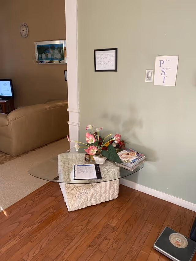 Glass-top table with a stone base holding a flower arrangement, magazines, and papers in a living room area with a couch and TV in the background.