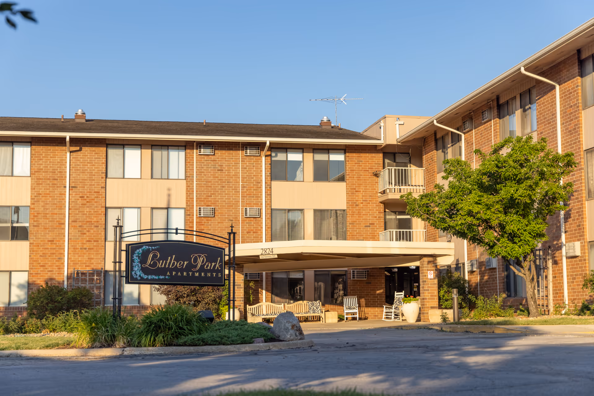 Exterior view of Luther Park Apartments, a three-story brick building with multiple windows. There is a covered entrance with benches and rocking chairs, a tree to the right, and a sign in front displaying the facility name.