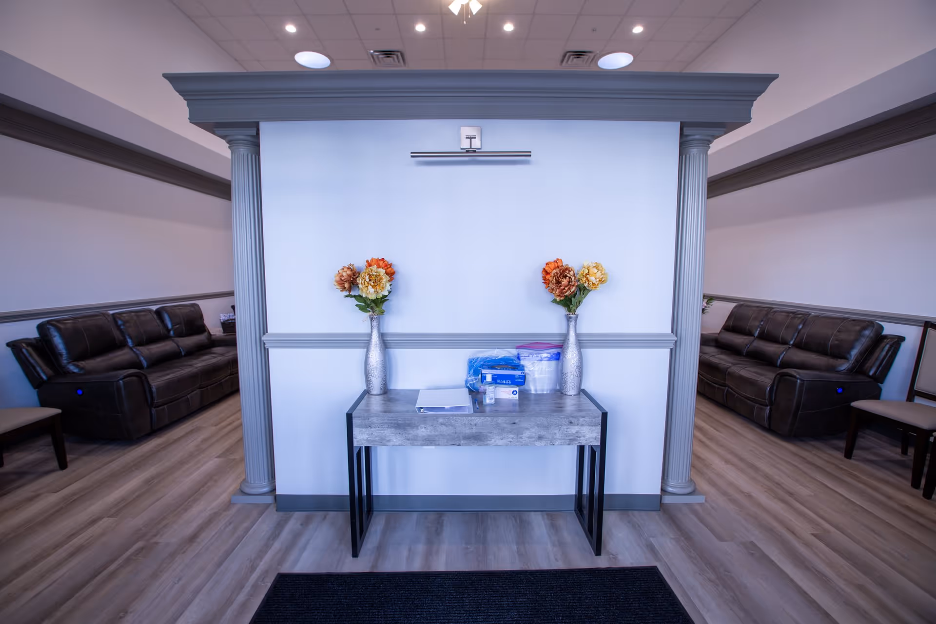 Interior view of a senior living facility lounge area with two brown leather sofas placed against opposite walls. A small table with two vases of artificial flowers and some supplies is positioned in the center against a white wall with decorative molding and columns on either side. The floor has light wood laminate flooring and the ceiling has recessed lighting and a ceiling fan.