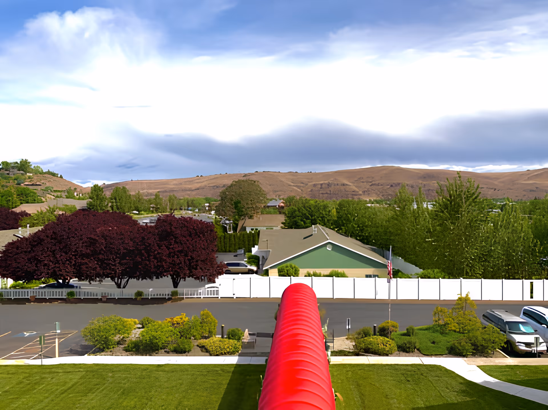 View over a red covered walkway toward a fenced residential street with trees, parked cars and rolling hills under a cloudy sky.