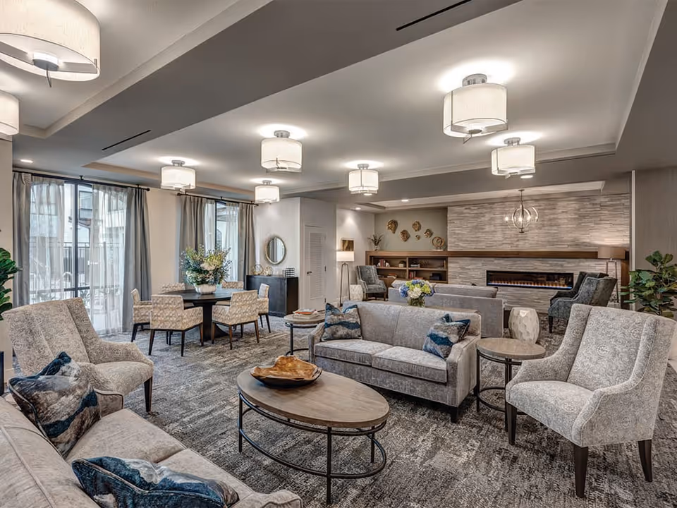 A spacious and well-lit living room area in a senior living facility featuring multiple gray upholstered sofas and armchairs arranged around wooden coffee tables. The room has large windows with sheer curtains, modern ceiling lights, a stone accent wall with a built-in fireplace, and decorative elements including plants, flowers, and wall art.